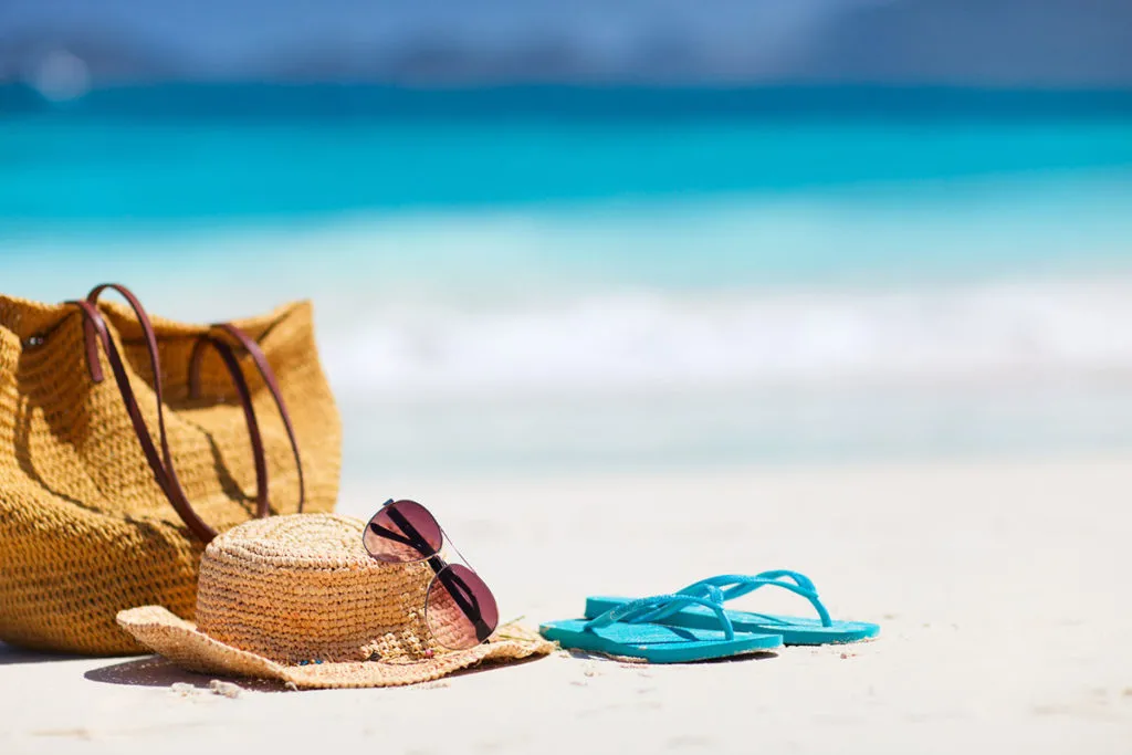 Straw hat, beach bag, and turquoise flip-flops on white sand near the ocean.
