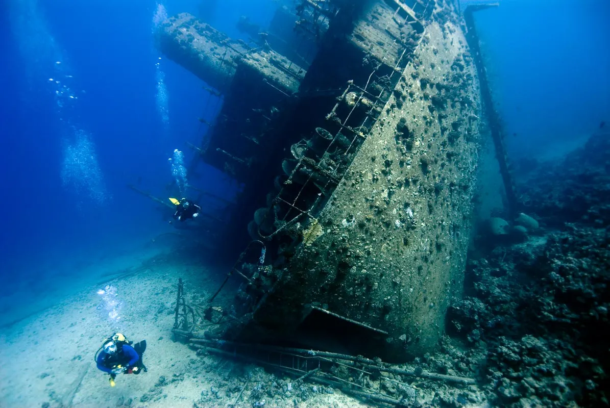 Scuba divers exploring the massive, algae-covered hull of the SS Thistlegorm shipwreck.